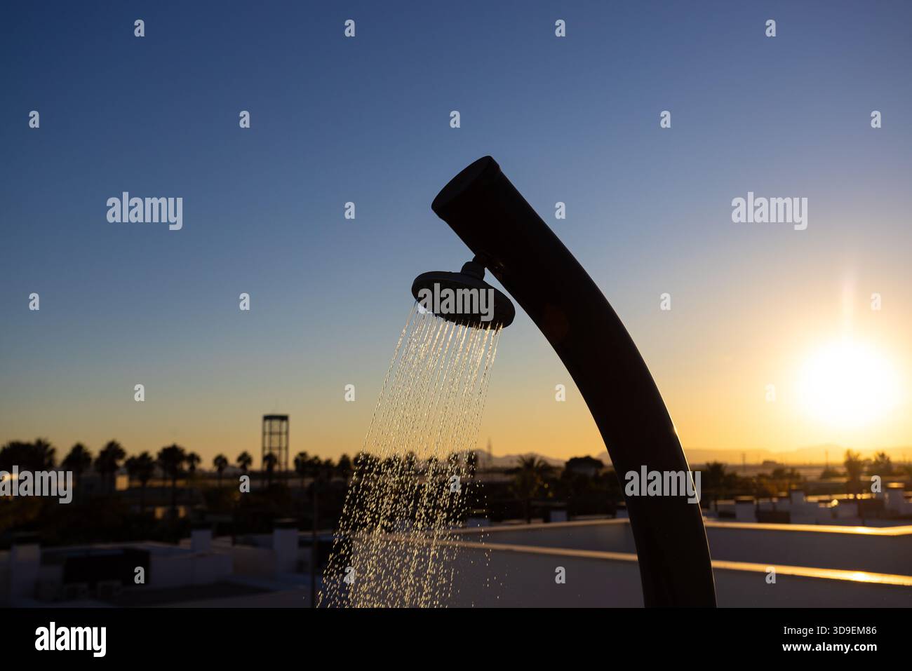 Die Dachdusche im Freien sprüht Wasser mit goldenem Sonnenlicht und palmengesäumter Stadtlandschaft in der Ferne. Stockfoto