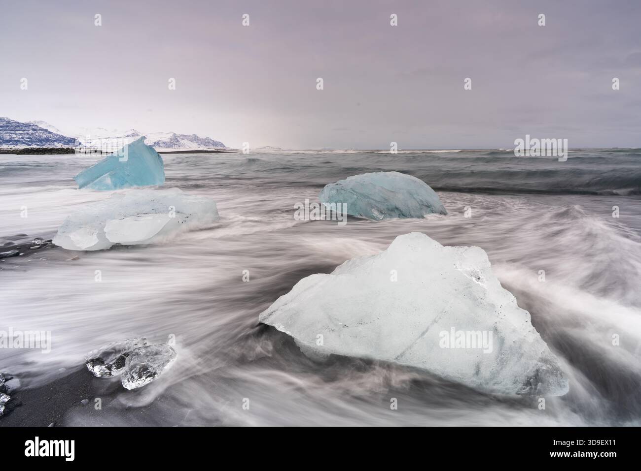 Blick auf lichtdurchlässige Eisberge, die an einem schwarzen Sandstrand verstreut sind und das turbulente Meer unter einem bewölkten Himmel treffen, Sveitarfelagid Hornafjordur, Sveitarfelagid Stockfoto