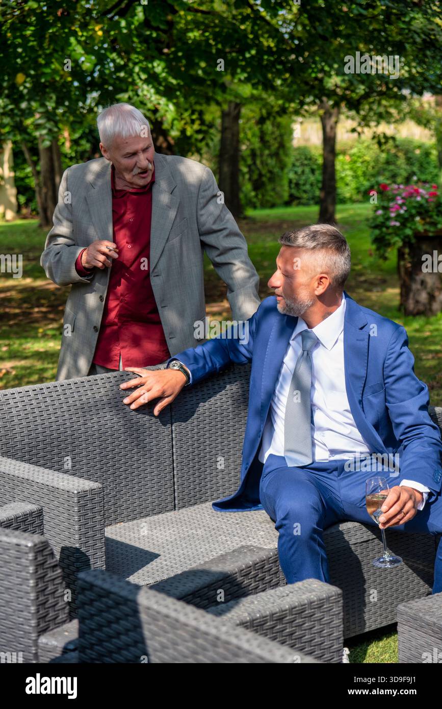 Freimütige Szene von zwei erwachsenen Männern, die sich während einer formellen Veranstaltung im Freien unterhalten. Ein Mann sitzt mit einem Glas Wein, der andere steht n Stockfoto