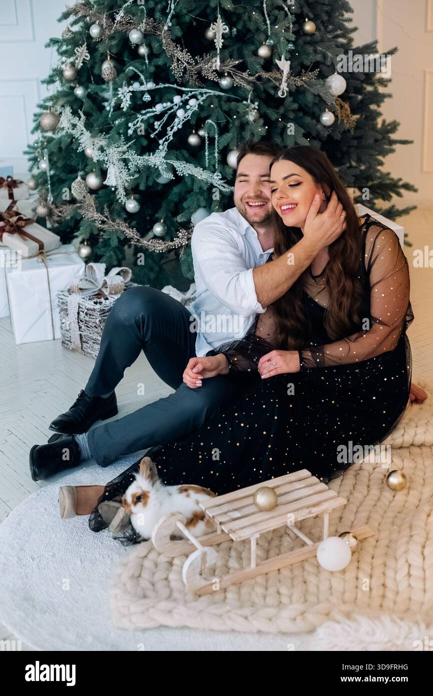Ein fröhliches Paar sitzt auf einem weichen Teppich neben einem wunderschön geschmückten Weihnachtsbaum. Sie teilen einen zärtlichen Moment, lächeln und spielen mit ihrem Haustier, Stockfoto