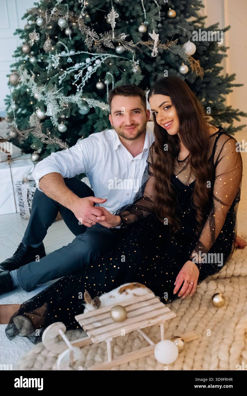 Ein Mann und eine Frau sitzen zusammen auf einem weichen Teppich neben einem wunderschön geschmückten Weihnachtsbaum. Sie teilen einen liebevollen Blick, gekleidet in stilvolle Urlaubskleidung, c Stockfoto
