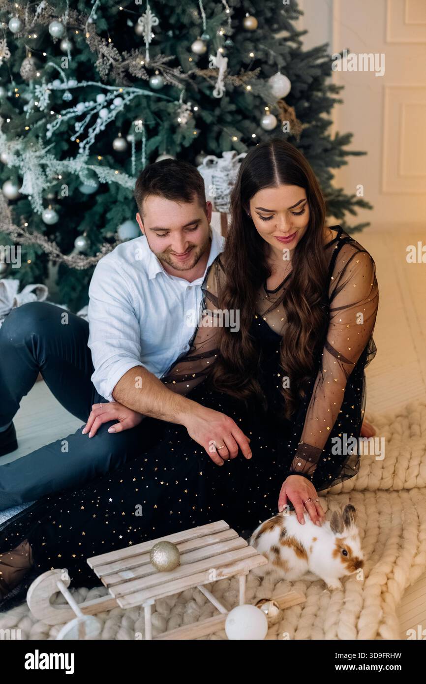 Ein Mann und eine Frau sitzen zusammen auf einem weichen Teppich und lächeln, während sie mit einem kleinen Kaninchen interagieren. Dahinter befindet sich ein wunderschön dekorierter Weihnachtstrier Stockfoto