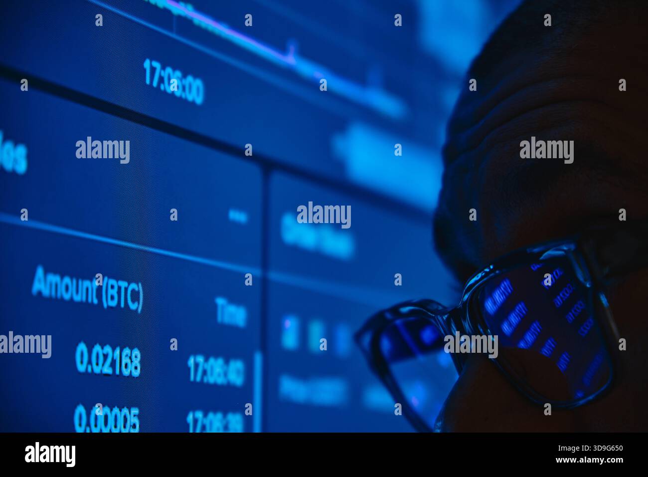 Ein Mann, der Live-Daten zum Handel mit Kryptowährungen auf einem blau beleuchteten Computermonitor beobachtet Stockfoto