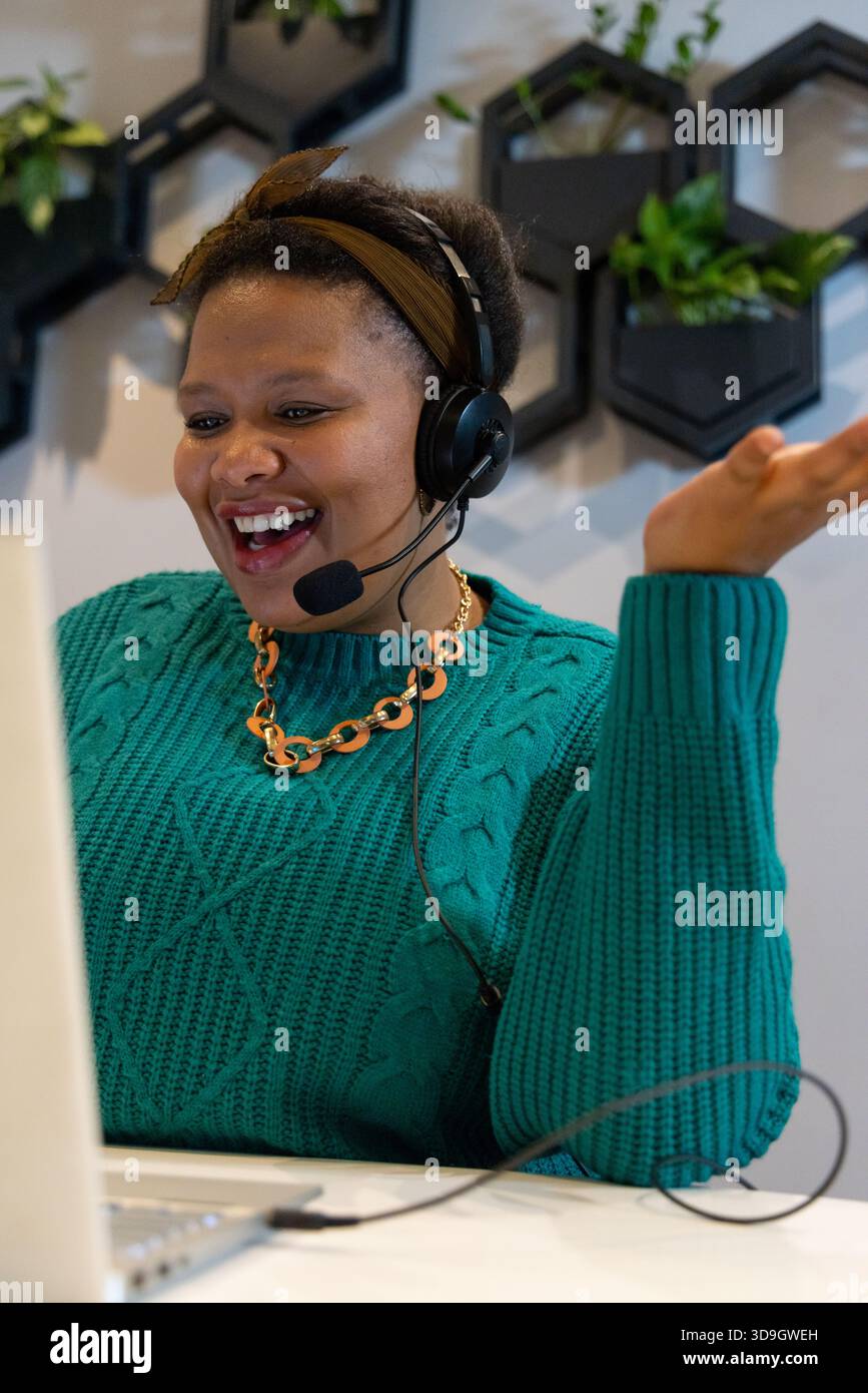 MittelErwachsene afroamerikanische Frau mit Gesten, Lächeln, Gespräch über Headset im Home Office mit Laptop Stockfoto