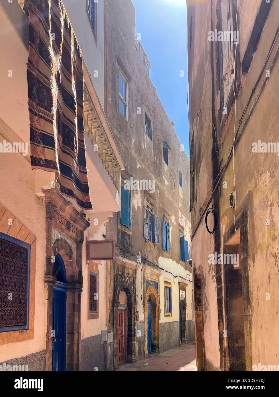 Schmale Gasse in Essaouira medina, Marokko. Verwitterte Steinfassaden, blaue Türen und Fensterläden, hängender Teppich, blauer Himmel, sonniger Tag. Stockfoto