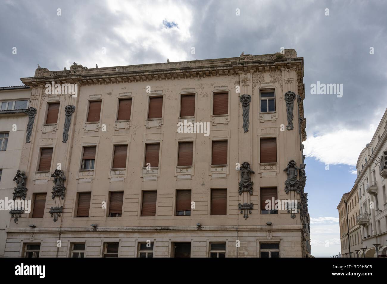 SPLIT, KROATIEN, 10.25.2025: Ein verwittertes, kunstvolles Gebäude säumt eine sonnenbeschienene Uferstraße. Skulpturale Reliefs zieren die Ecke, während geschlossene Fensterläden Hi Stockfoto