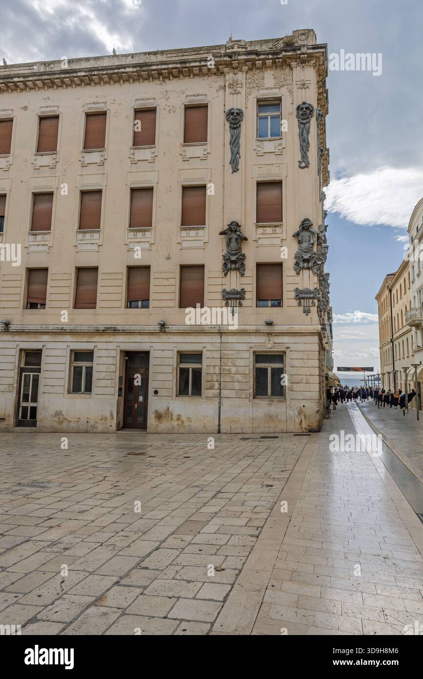 SPLIT, KROATIEN, 10.25.2025: Ein verwittertes, kunstvolles Gebäude säumt eine sonnenbeschienene Uferstraße. Skulpturale Reliefs zieren die Ecke, während geschlossene Fensterläden Hi Stockfoto