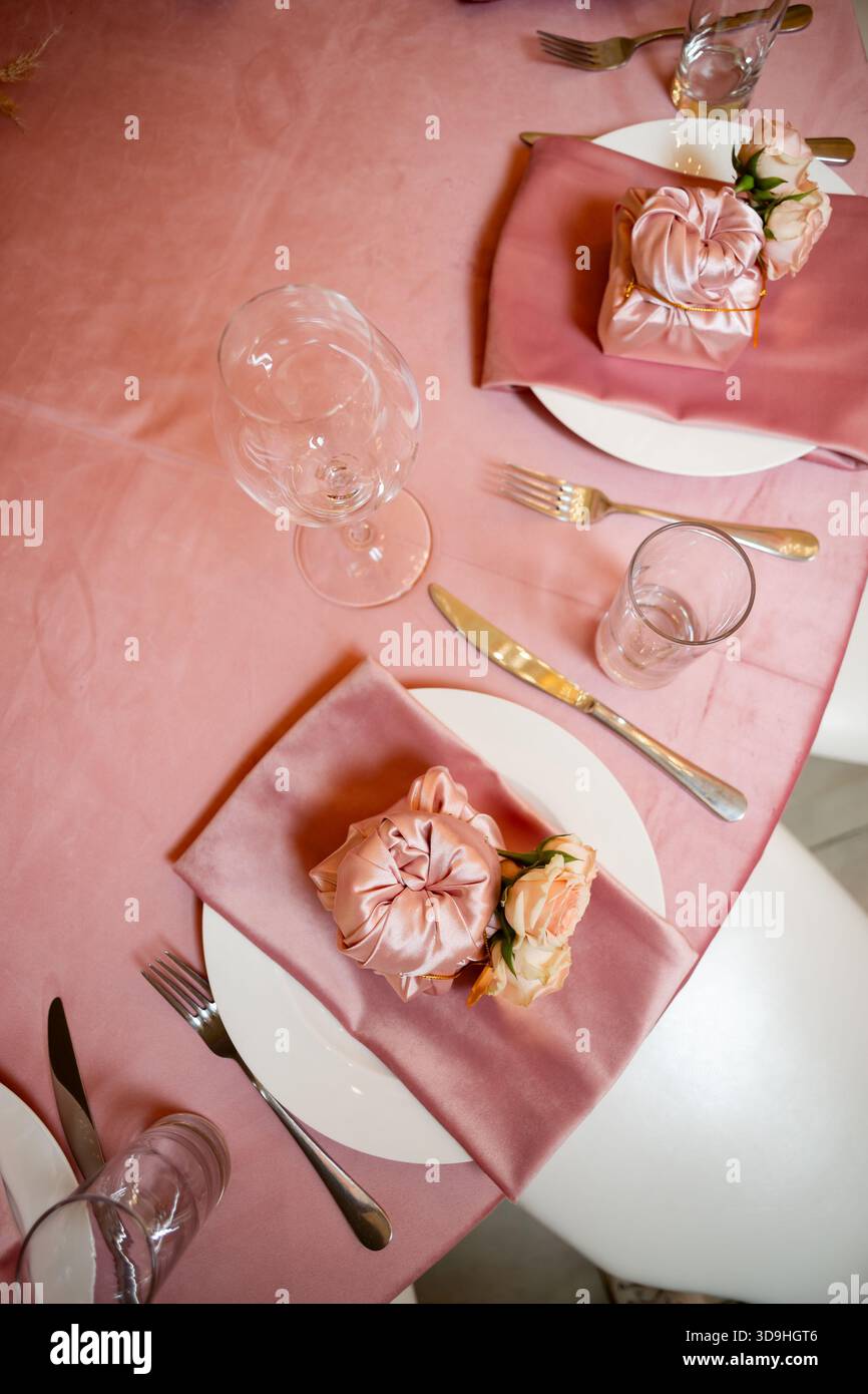 Das Foto zeigt eine Ansicht von oben nach unten auf einen rosafarbenen Tisch mit Satin-Servietten, zarten Rosen, einer Geschenkbox und ordentlich angeordnetem Besteck. Stockfoto