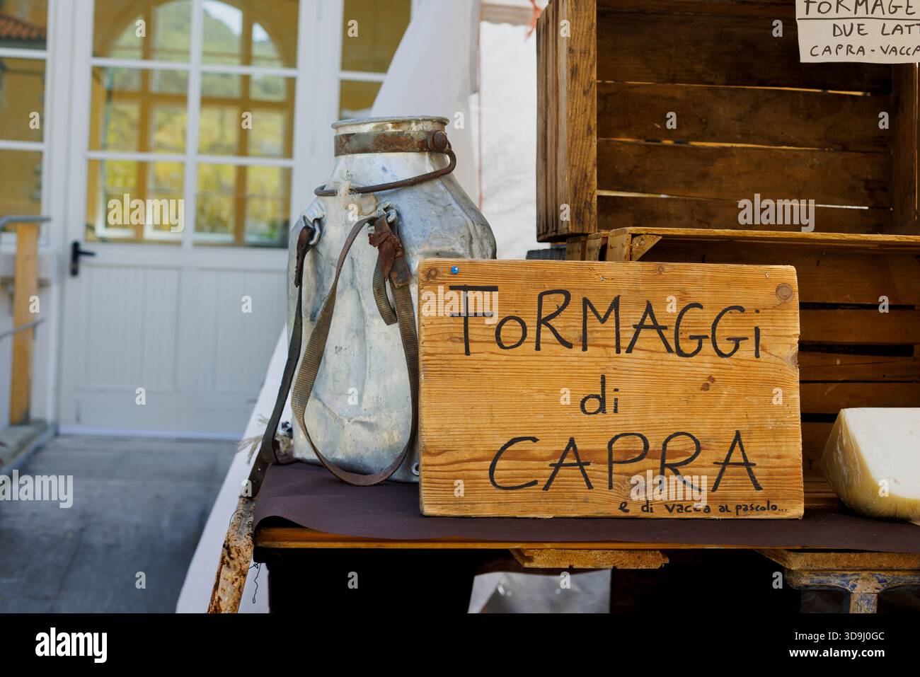 Hölzerne Schilder, die Ziegenkäse zum Verkauf anbieten, auf einem rustikalen Tisch im Freien mit einem alten Aluminium-Milchtransportbehälter ausgestellt. Ruft „Farm-to-tab“ hervor Stockfoto