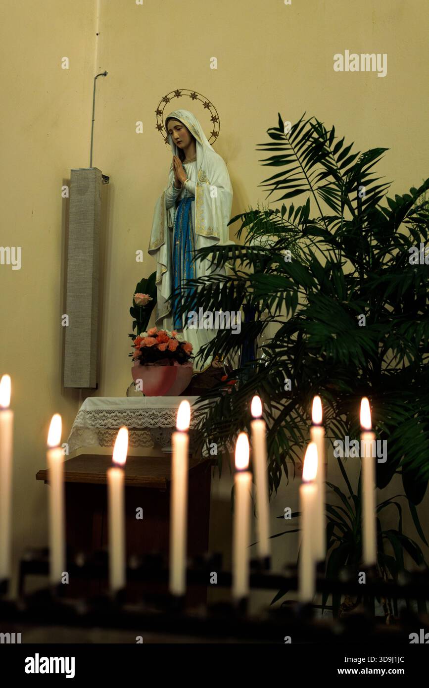 Kircheninnenraum mit einer Statue der Madonna im Hintergrund, in selektiver Fokussierung dargestellt. Im Vordergrund zündete Votivkerzen zum Opferopfer an Stockfoto