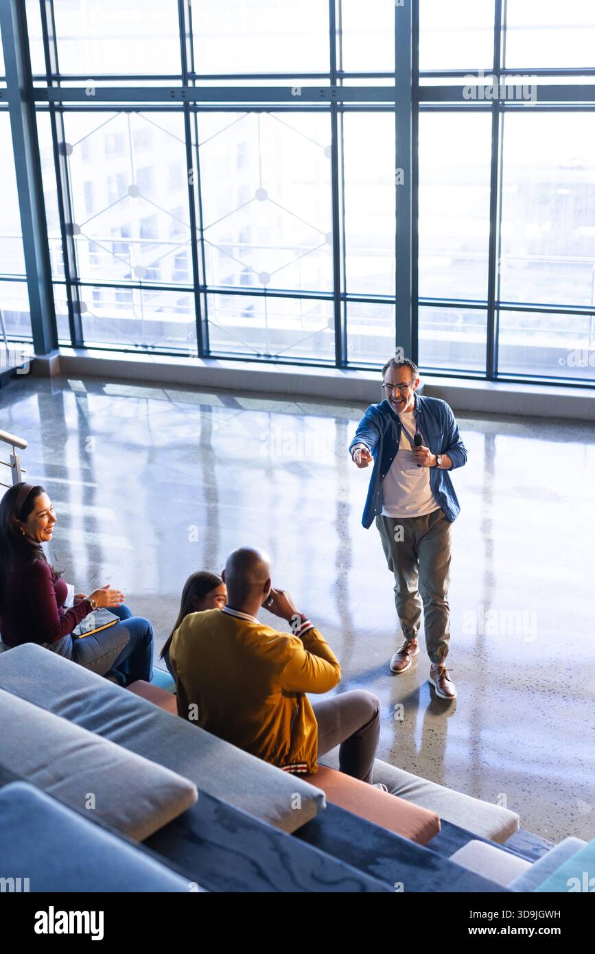 Verschiedene Kollegen, die an einem Workshop im Atrium teilnehmen, und Moderatoren, die Gesten mit Mikrofon in der Nähe von mehrstufigen Sitzen halten Stockfoto