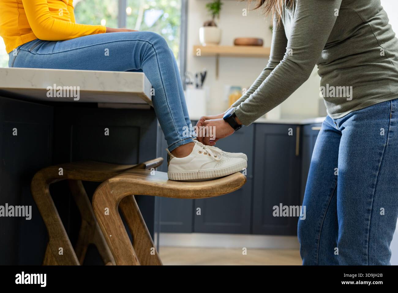 Mutter fesselt den weißen Sneaker der Tochter auf hölzernem Barhocker bei Marmorküche mit Smartwatch Stockfoto
