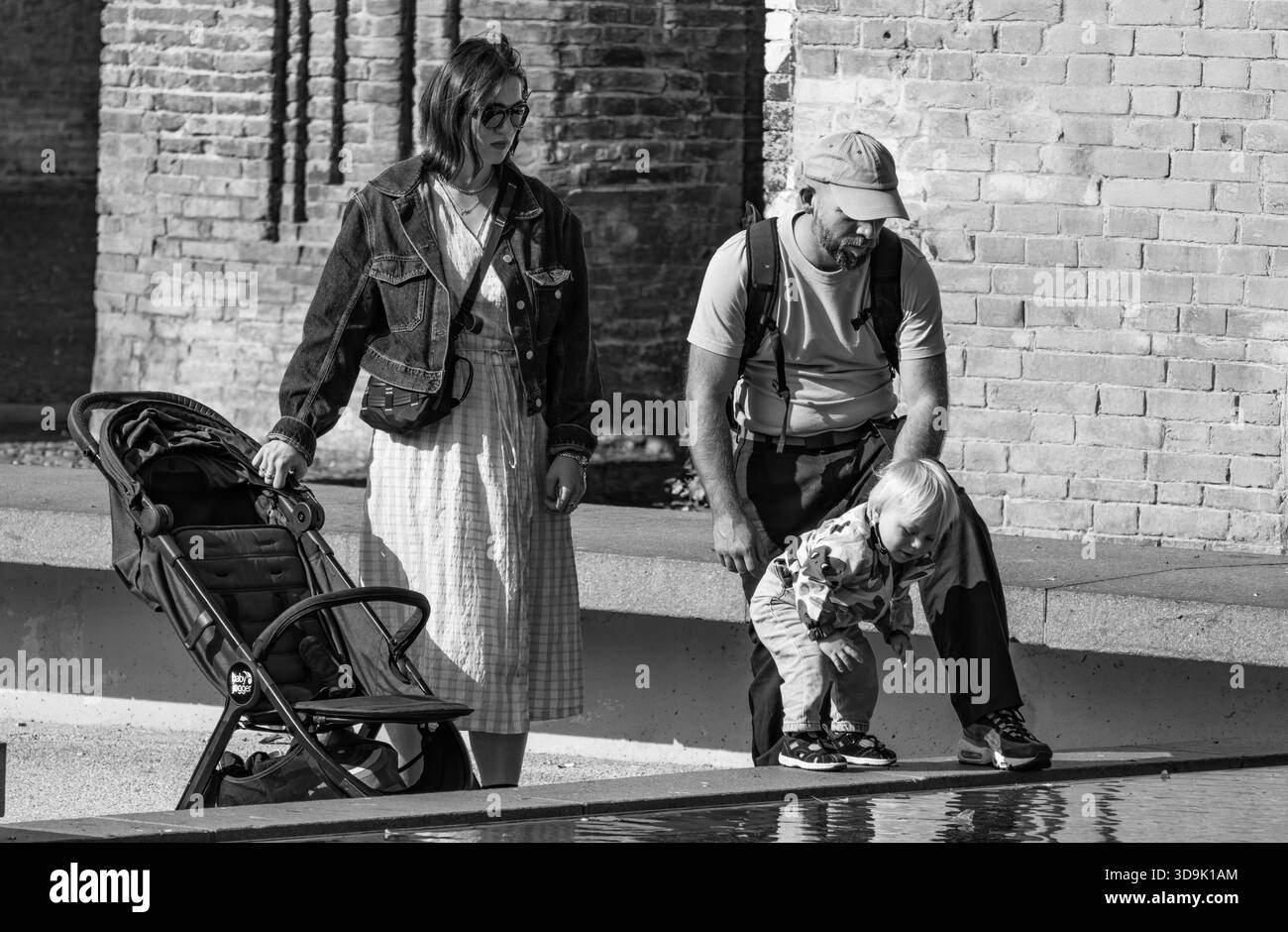Eine zeitlose Szene, in der sich ein Kind zum Wasser beugt, geführt von geduldigen Eltern, eingerahmt von alten Mauern. Stockfoto