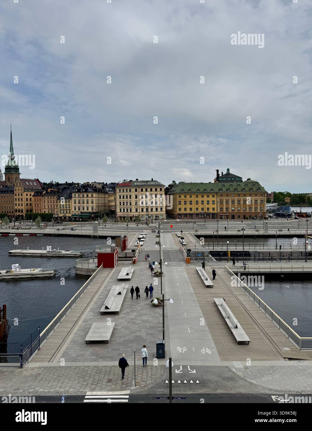 Fußgänger- und Radbrücke am Ufer im Zentrum von Stockholm, Schweden, mit Menschen, die zu den historischen Gebäuden von Gamla Stan laufen. - Smartphone-aufgenommenes Stockfoto