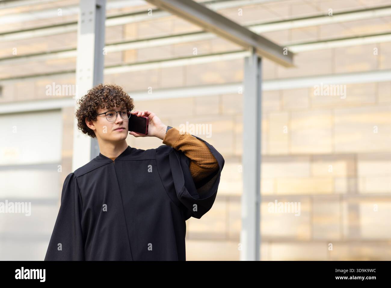 Männlicher Absolvent im Kleid, der unter einer Pergola aus Metall in der Nähe einer holzgetäfelten Wand steht und das Smartphone am Ohr hält Stockfoto
