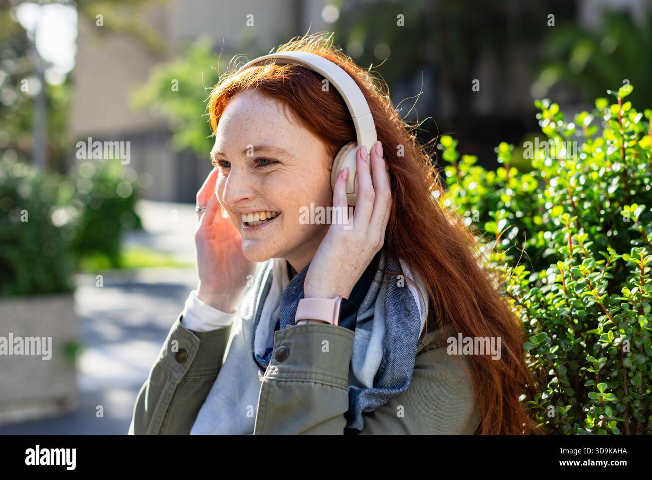 Rothaarige Frau, die über-Ohr-Kopfhörer hält und Smartwatch im Innenhof überprüft Stockfoto