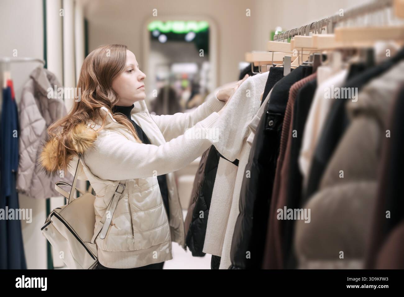 Eine schöne 30- bis 35-jährige Frau in einer weißen Jacke wählt und pflegt Kleidung in den Regalen und zieht Kleidung aus dem Kleiderbügel Stockfoto