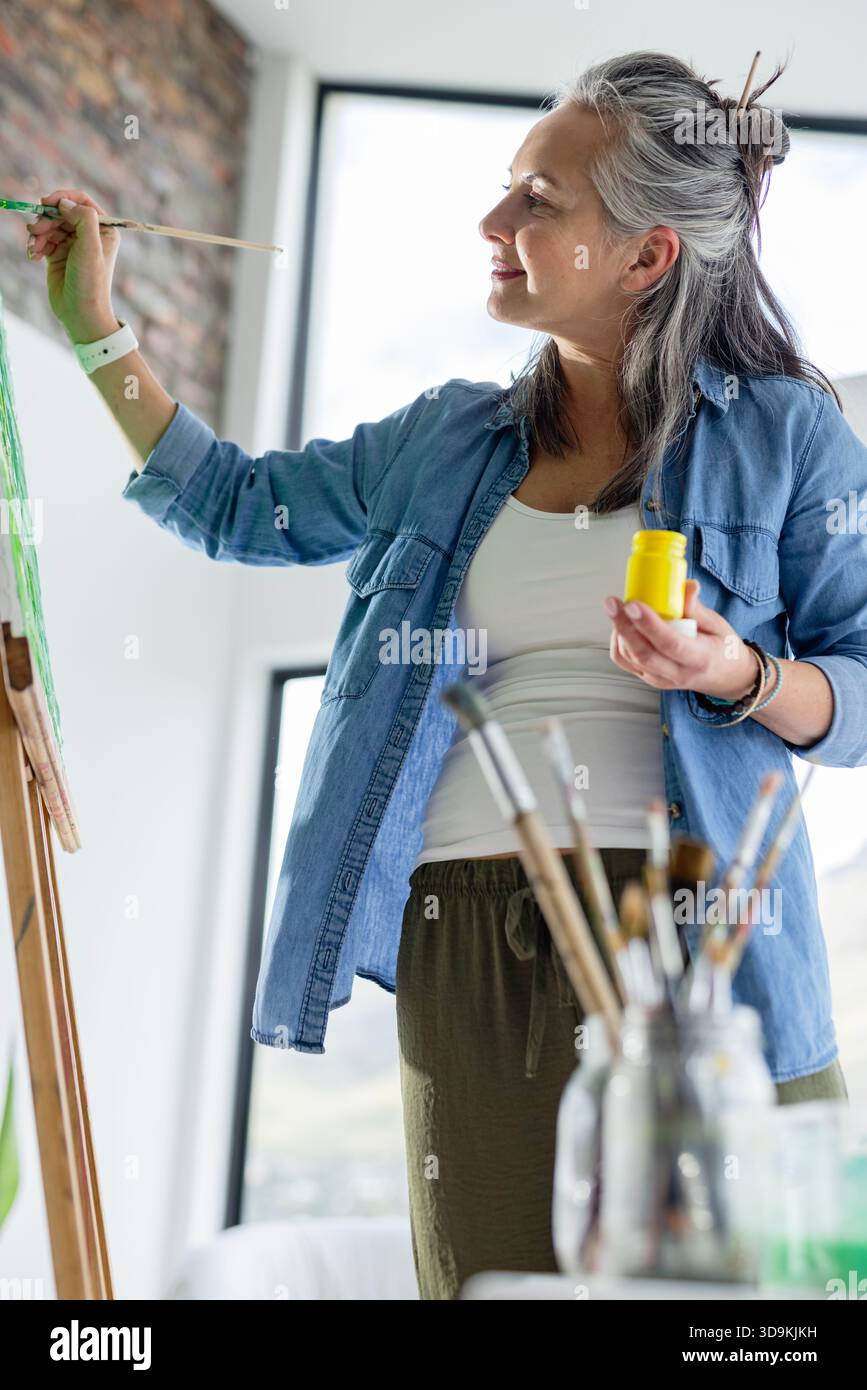 Maler mit Pinsel und gelbem Farbbehälter, der grüne Farbe auf Leinwand auf Staffelei im Studio aufträgt Stockfoto