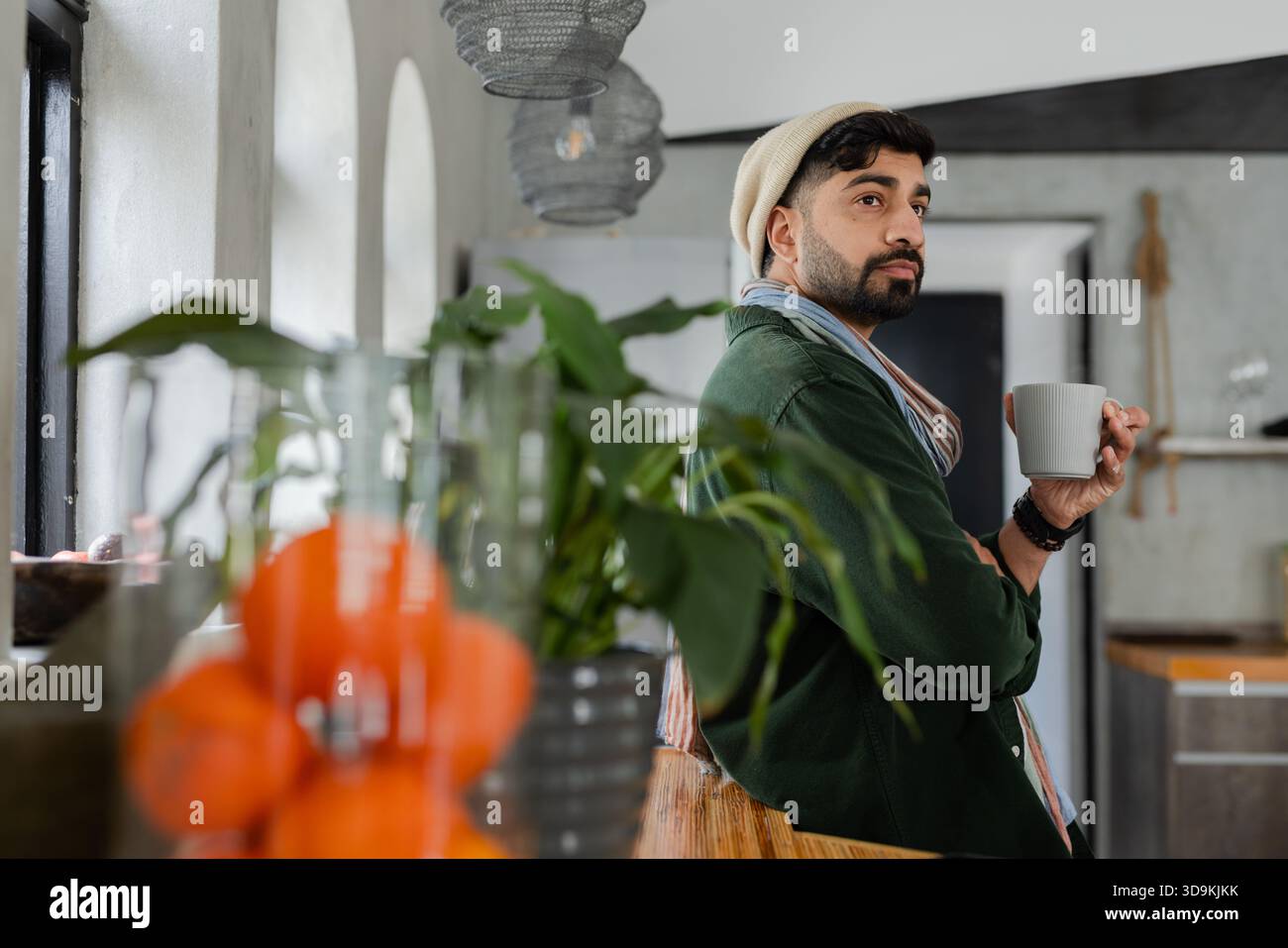 Indischer Mann lehnt sich an eine Holztheke in der Küche und hält einen weißen Becher mit Strickmütze, Kopierraum Stockfoto