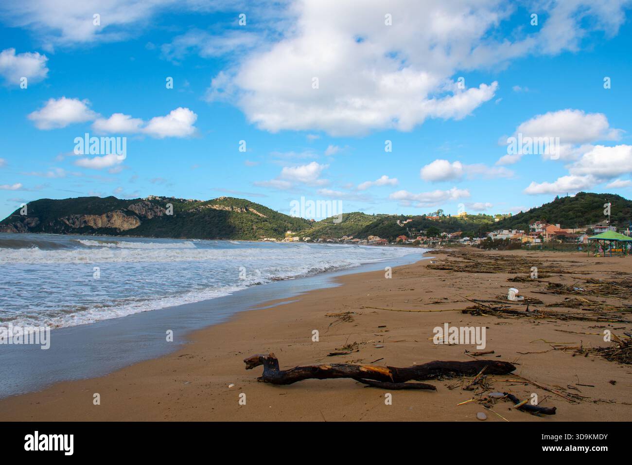 Welliges Meer am Sandstrand Agios Georgios Pagon, Korfu, Griechenland, mit grünen Hügeln und Häusern in der Ferne unter blauem Himmel Stockfoto