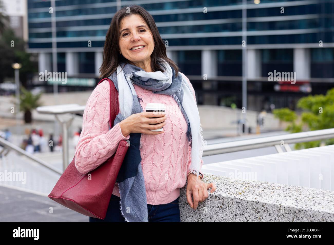 Frau, die sich auf dem Fußweg mit Blick auf den City plaza lehnte, trug einen Schal mit einer Kaffeetasse und einer roten Tragetasche Stockfoto