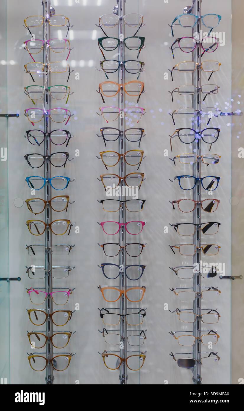 Das Foto zeigt Dutzende von Brillenrahmen in verschiedenen Farben und Formen, die ordentlich auf einem vertikalen Displaygestell in einem optischen Geschäft angeordnet sind. Stockfoto