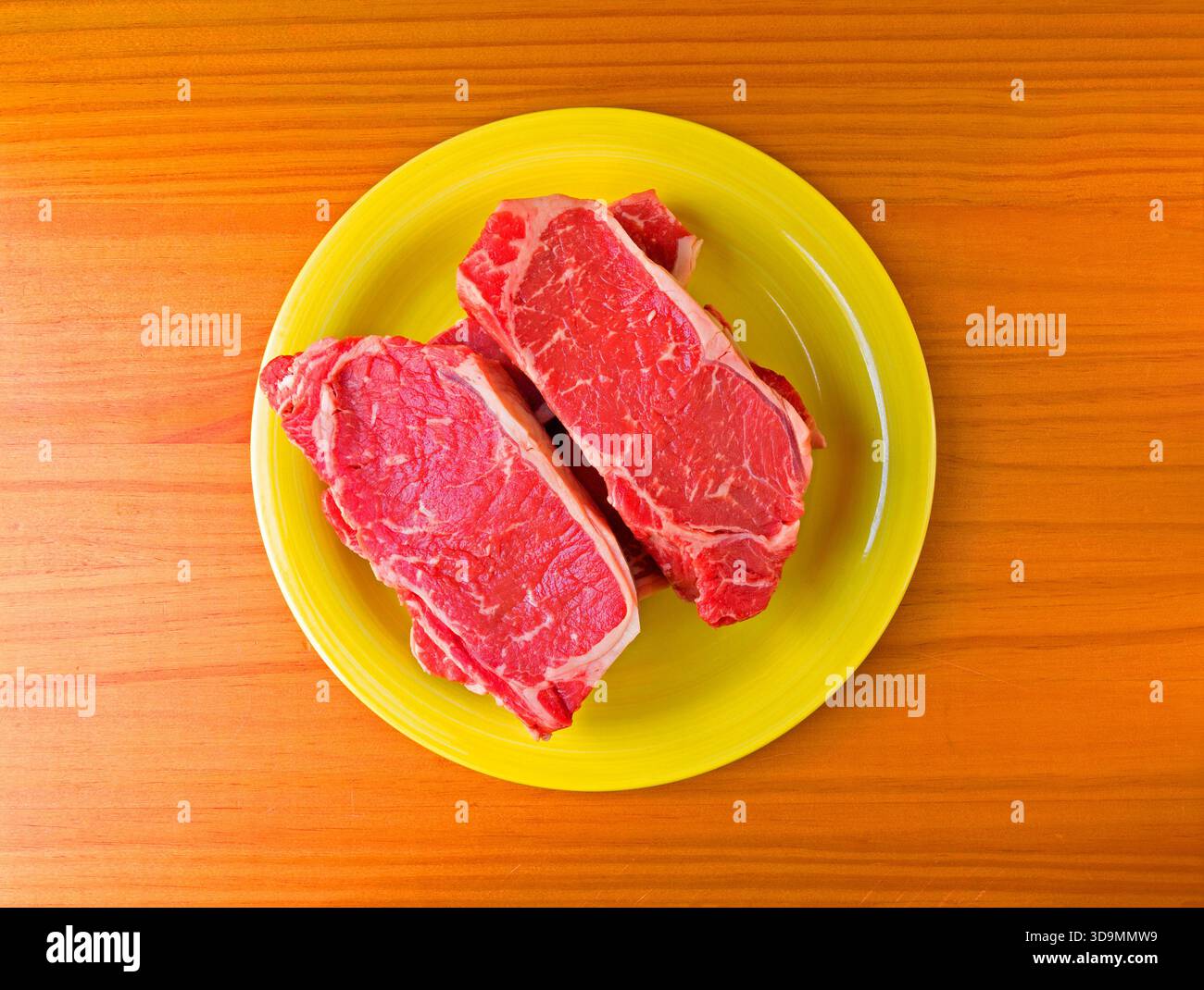 Blick von oben auf die Steaks des New Yorker Streifens auf einer gelben Platte auf einer mit Tageslicht beleuchteten Holzplatte. Stockfoto