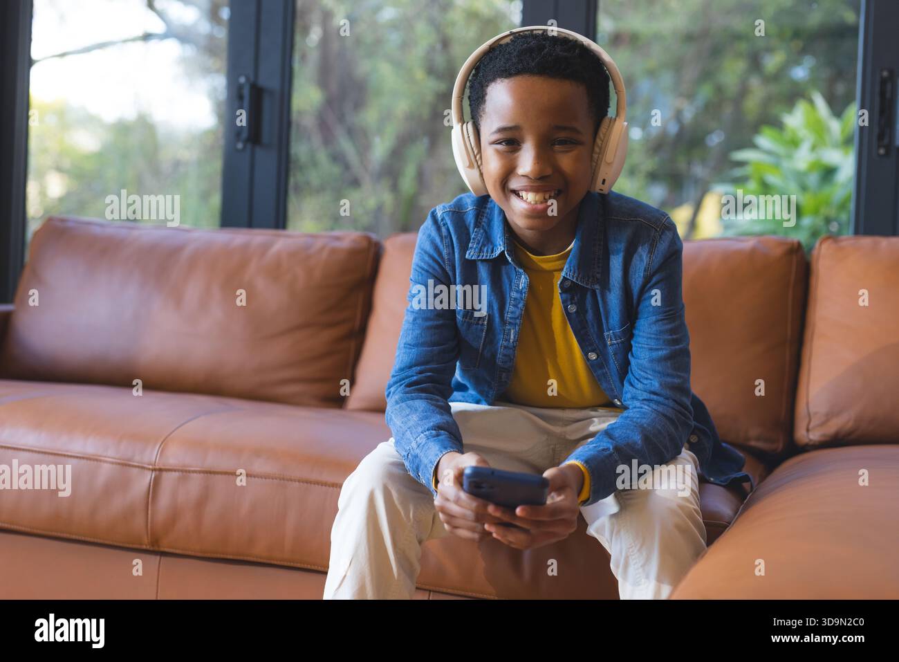 Afroamerikanischer Junge lächelt, während er das Smartphone im Wohnzimmer mit weißen Kopfhörern auf dem Sofa hält Stockfoto