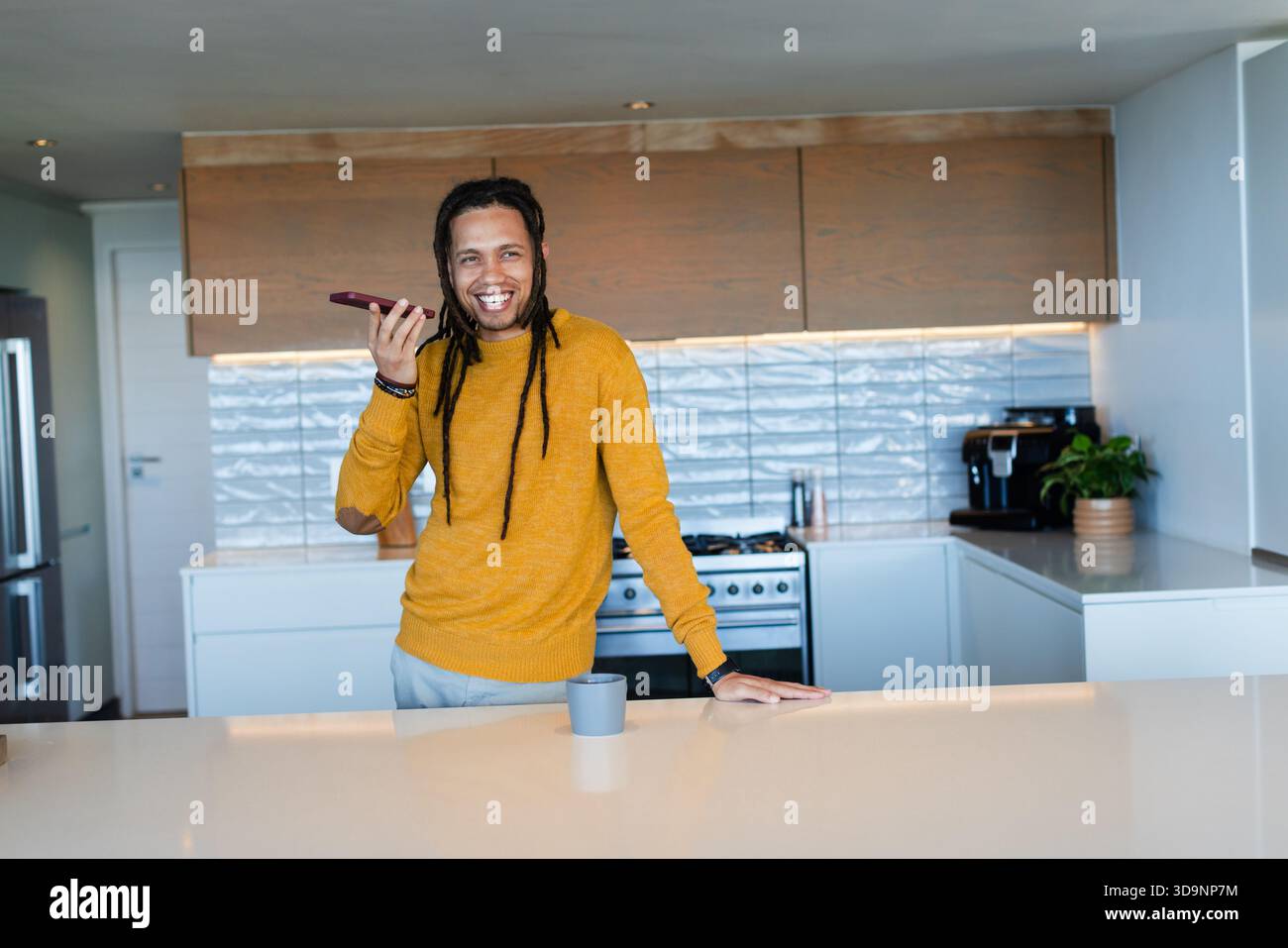 Afroamerikanischer Mann, der sich in das Smartphone lehnt, während er sich auf die Theke in der Küche mit Kaffeetasse lehnt Stockfoto