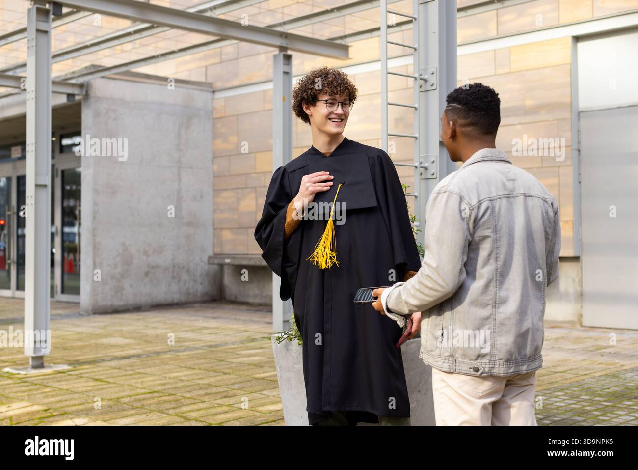Verschiedene männliche Freunde im Kleid zeigen auf die Quaste und benutzen das Smartphone neben Pflanzgefäßen auf dem Campus Quad Stockfoto
