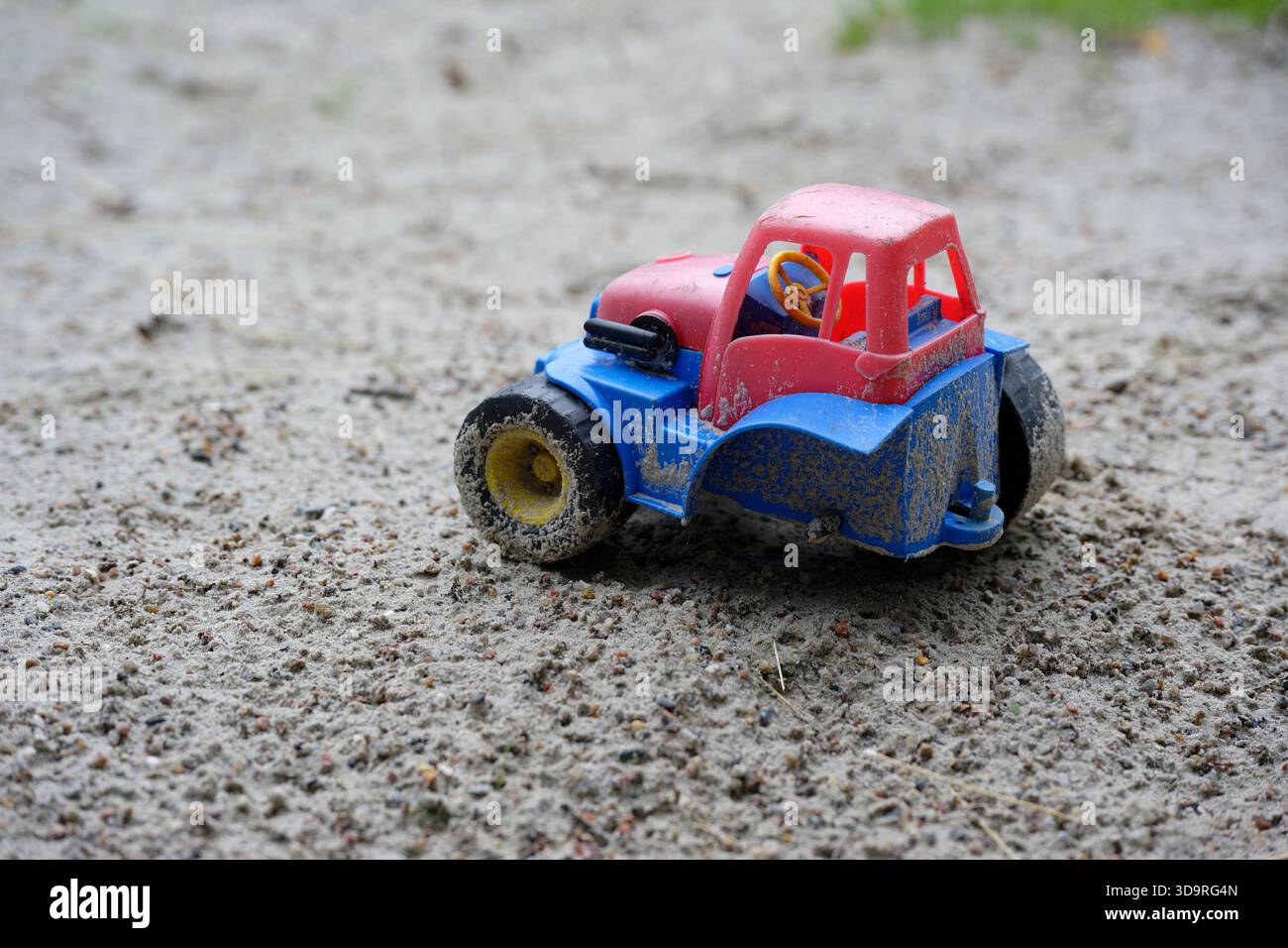 Im Sand eines Sandkastens befindet sich ein blaurotes Spielzeug, ein Traktor mit einem großen Hinterrad fehlt Stockfoto