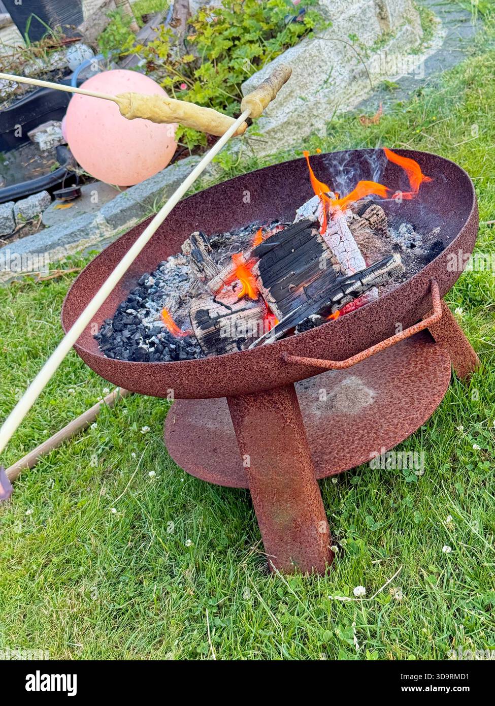 Eine große rostige Feuerstelle auf drei Beinen steht auf Gras mit brennenden Brettern. Die Leute braten Teig auf Stöcken über den Flammen. Die einfache Einrichtung fühlt sich an Stockfoto