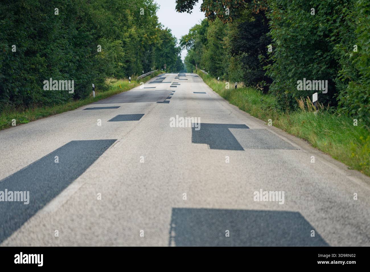 Ein ländlicher Abschnitt verläuft zwischen Bäumen, wo unzählige Stellen den hellen Asphalt markieren. Dunkles Bitumen füllt alle paar Meter alte Schlaglöcher. Die gemusterten s Stockfoto