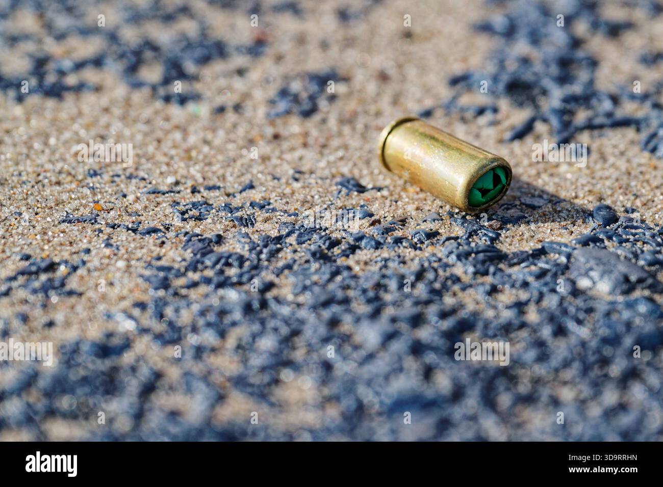 Dunkle Kieselsteine verstreuen sich über hellen Sand, ein verbrauchter Kugelmantel liegt unter ihnen, das Messing fängt die Sonne ein, der Kontrast fühlt sich scharf und stark an, der fram Stockfoto