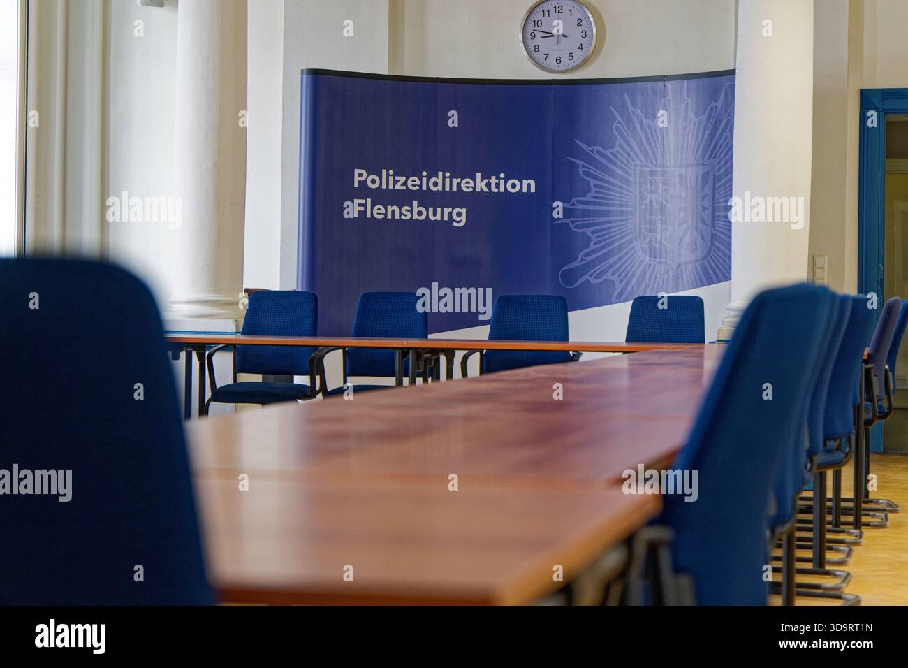 Lange Tische säumen einen hellen Saal mit blauen Stühlen, auf dem Schild steht Polizei Direktion Flensburg, Uhren markieren die Zeit, weiße Wände und Säulen halten den Sprung Stockfoto