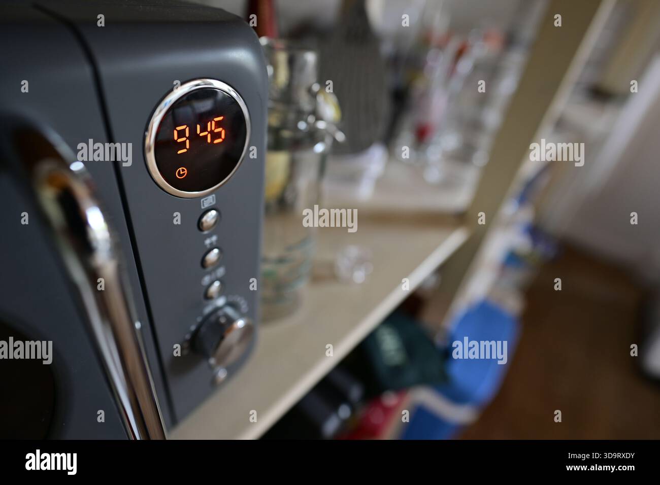 Eine Nahaufnahme eines Mikrowellenherds, die die Zeit 9:45 anzeigt. Der Hintergrund ist mit verschiedenen Küchenartikeln und Gläsern versehen, die eine gemütliche Küchenatmosphäre schaffen Stockfoto
