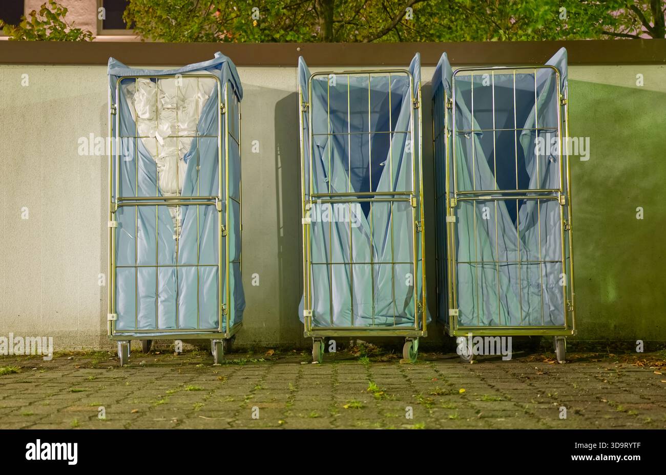 Drei leere Wäschewagen aus einem Hotel mit blauen Abdeckungen, die an einer Wand in einem schwach beleuchteten Bereich stehen. Die Wagen stehen auf einem Kopfsteinpflaster Stockfoto