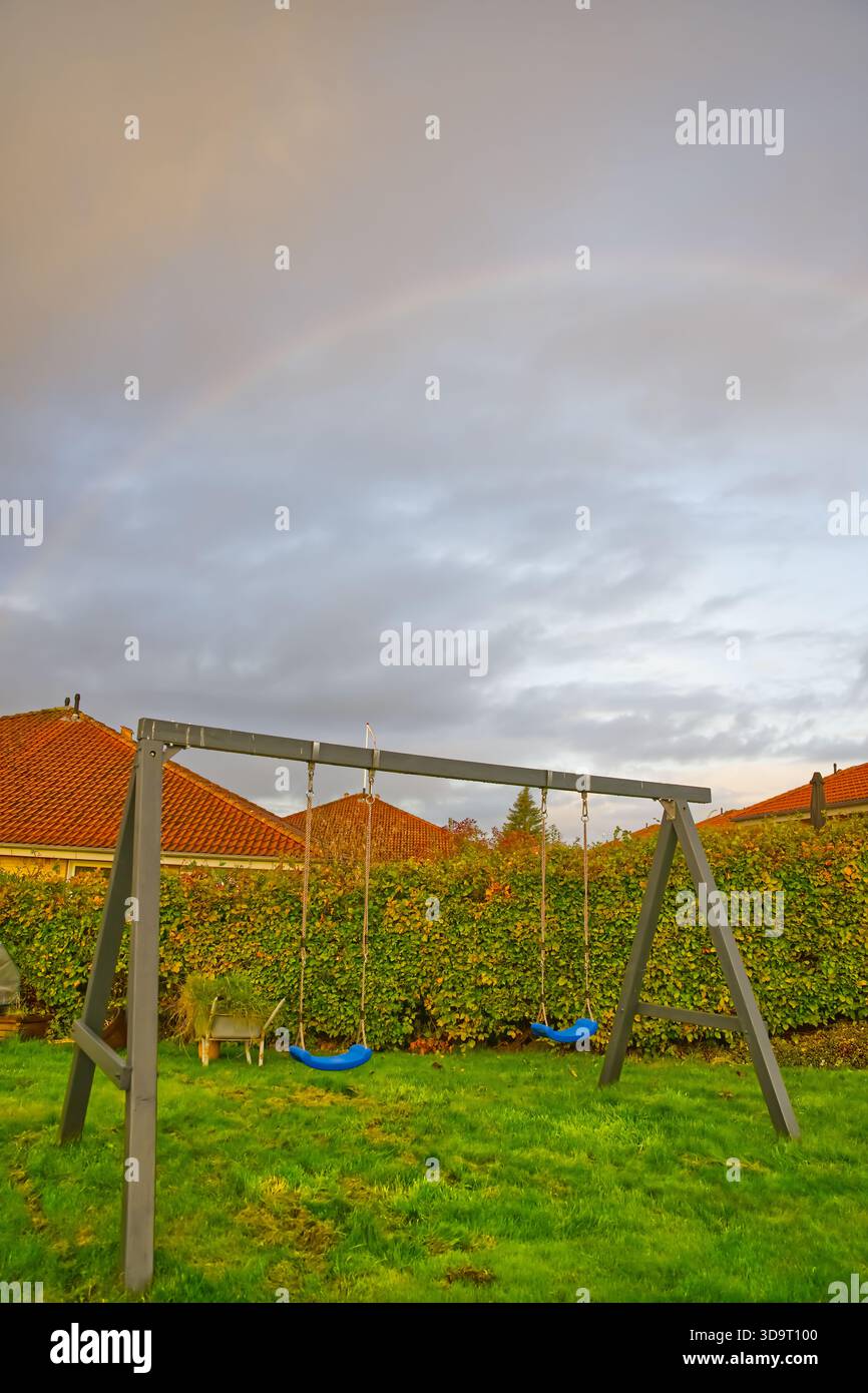 Eine Spielplatzschaukel mit drei Schaukeln in einem grasbewachsenen Gebiet, umgeben von einer Hecke und Wohnhäusern im Hintergrund unter einem bewölkten Himmel. Stockfoto