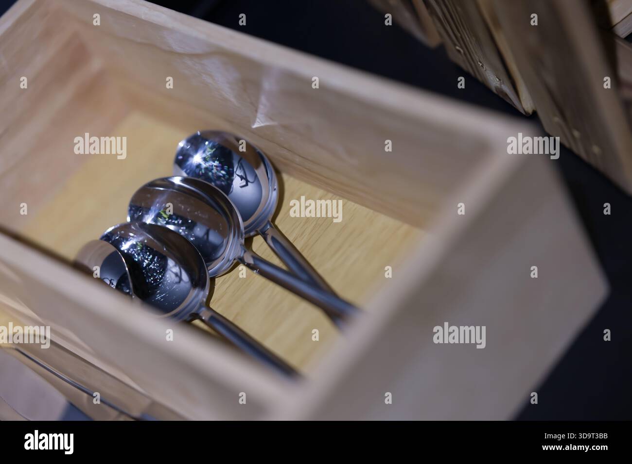 Eine Holzkiste mit drei glänzenden Edelstahllöffeln, die Licht und Umgebung reflektieren. Die Box hat ein schlichtes Design, das die Utensilien i betont Stockfoto
