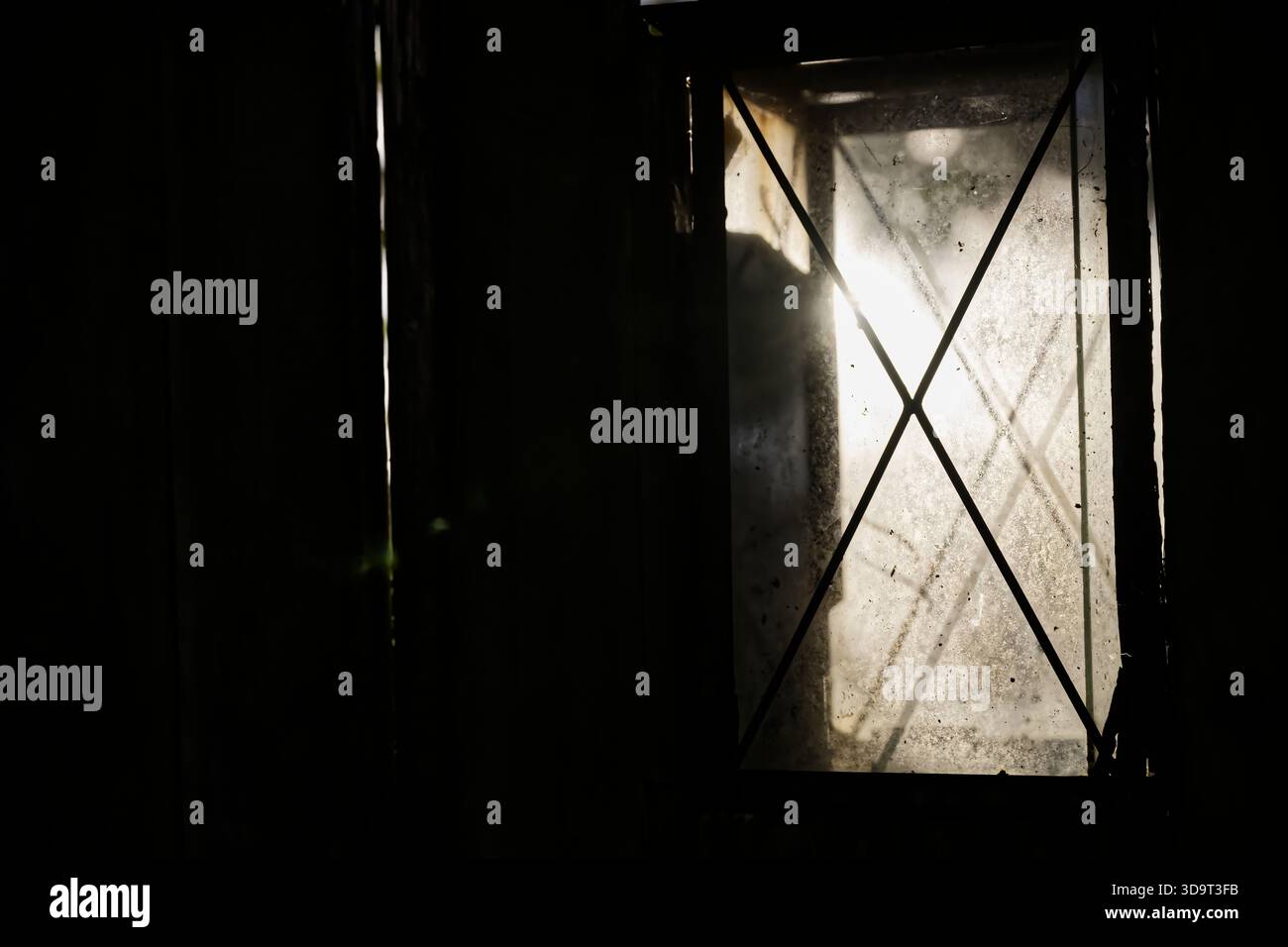 Ein schwach beleuchtetes Fenster mit kreuzförmigem Muster, das teilweise von Schatten verdeckt ist, sodass eine kleine Menge Licht durchdringen kann. Stockfoto