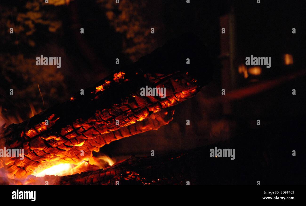 Eine Nahaufnahme eines brennenden Holzschlauchs am Lagerfeuer mit glühender Glut und Flammen vor dunklem Hintergrund, die eine warme und gemütliche Atmosphäre schaffen. Stockfoto