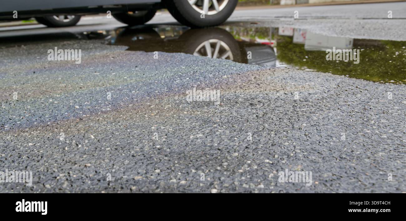 Eine Nahaufnahme einer nassen Asphaltstraße mit einer Pfütze, die einen Autoreifen reflektiert, und farbenfrohen Ölleckenmustern auf der Oberfläche, die durch ein Ölleck des Fahrzeugs verursacht wurden Stockfoto