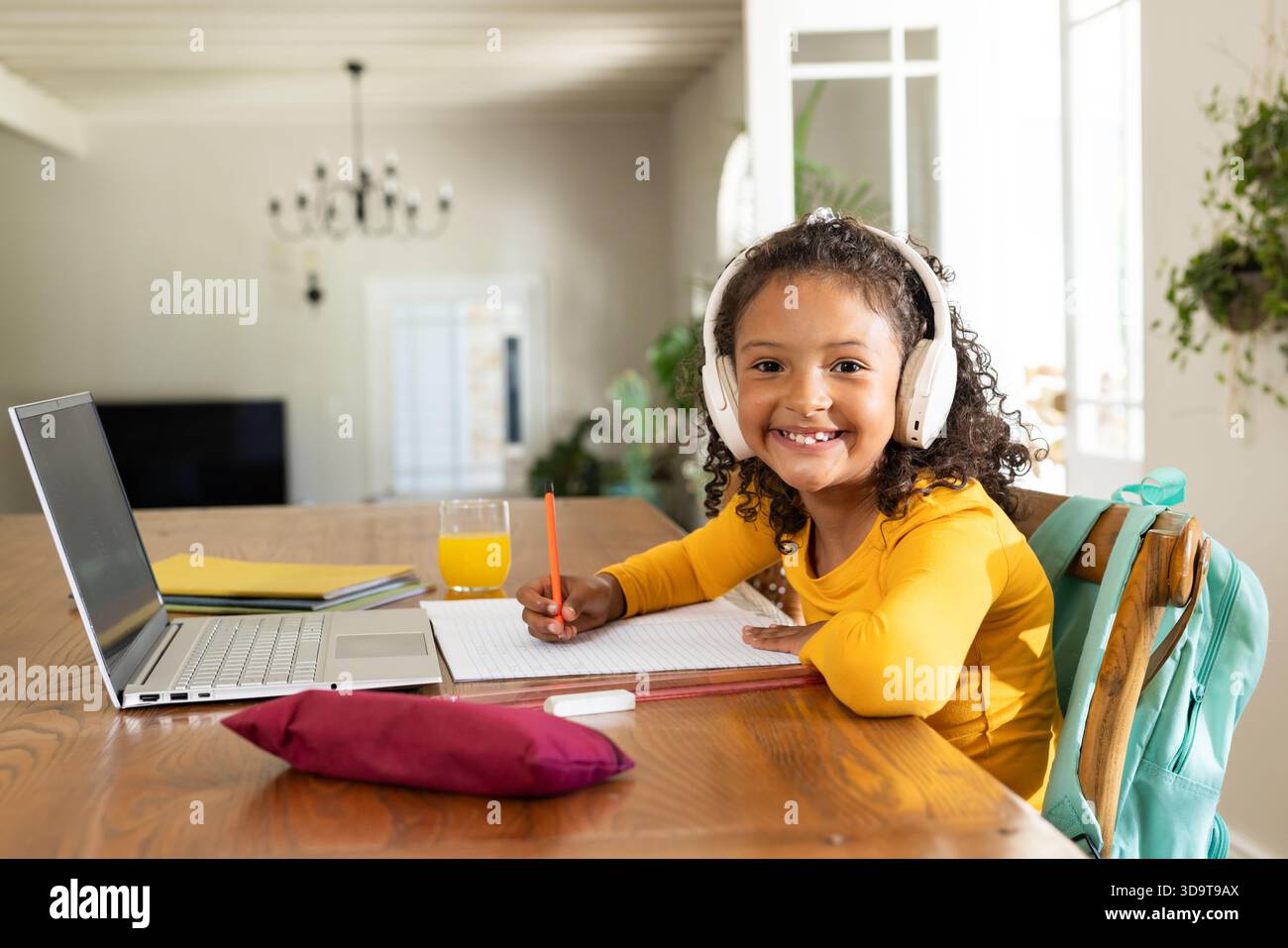 Afroamerikanisches Mädchen, das Kopfhörer trägt, sitzt am Tisch zu Hause und schreibt in Notebook mit Laptop Stockfoto