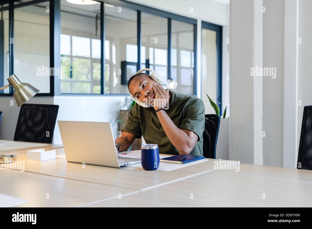 Afroamerikanischer Mann, der am Schreibtisch sitzt und Kopfhörer mit Laptop und Reisebecher trägt Stockfoto