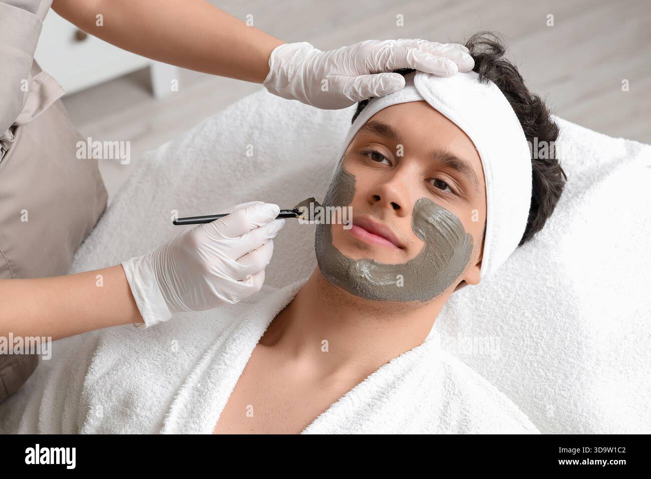 Kosmetikerin, die im Schönheitssalon Tonmaske auf das Gesicht des Mannes aufträgt Stockfoto
