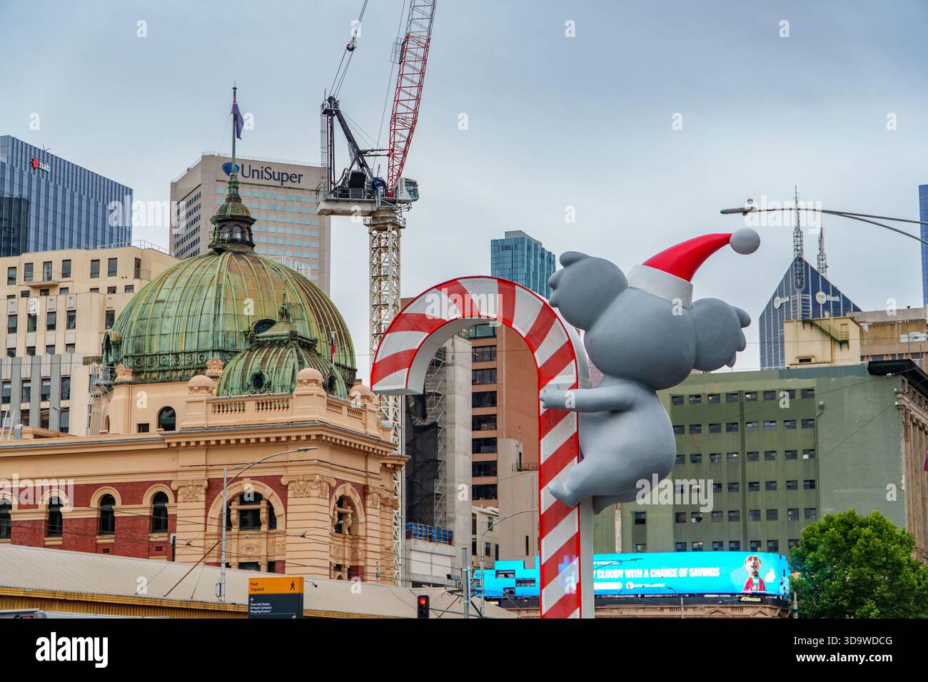 Ein grauer Koala im Weihnachtsmann-Hut hängt an einem riesigen Zuckerrohr, mit Melbournes Flinders Street Station und Wolkenkratzern unter einem bewölkten Himmel. Stockfoto