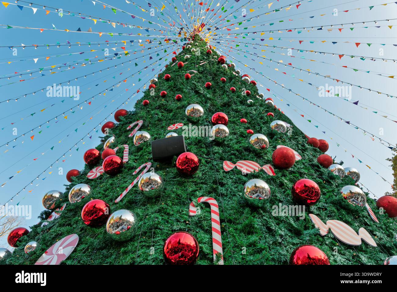 Riesiger, hell dekorierter Weihnachtsbaum im Freien, von unten betrachtet, mit roten und silbernen Ornamenten und festlichen Wimpelschnüren vor blauem Himmel. Stockfoto