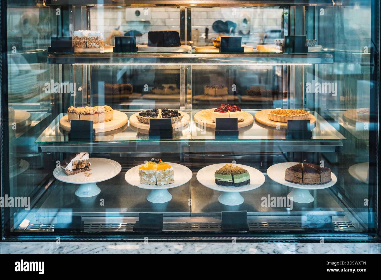 Verschiedene Kuchen, Torten und Desserts auf Tellern und Holztabletts in einer gekühlten Glasvitrine in der Bäckerei oder im Café Stockfoto