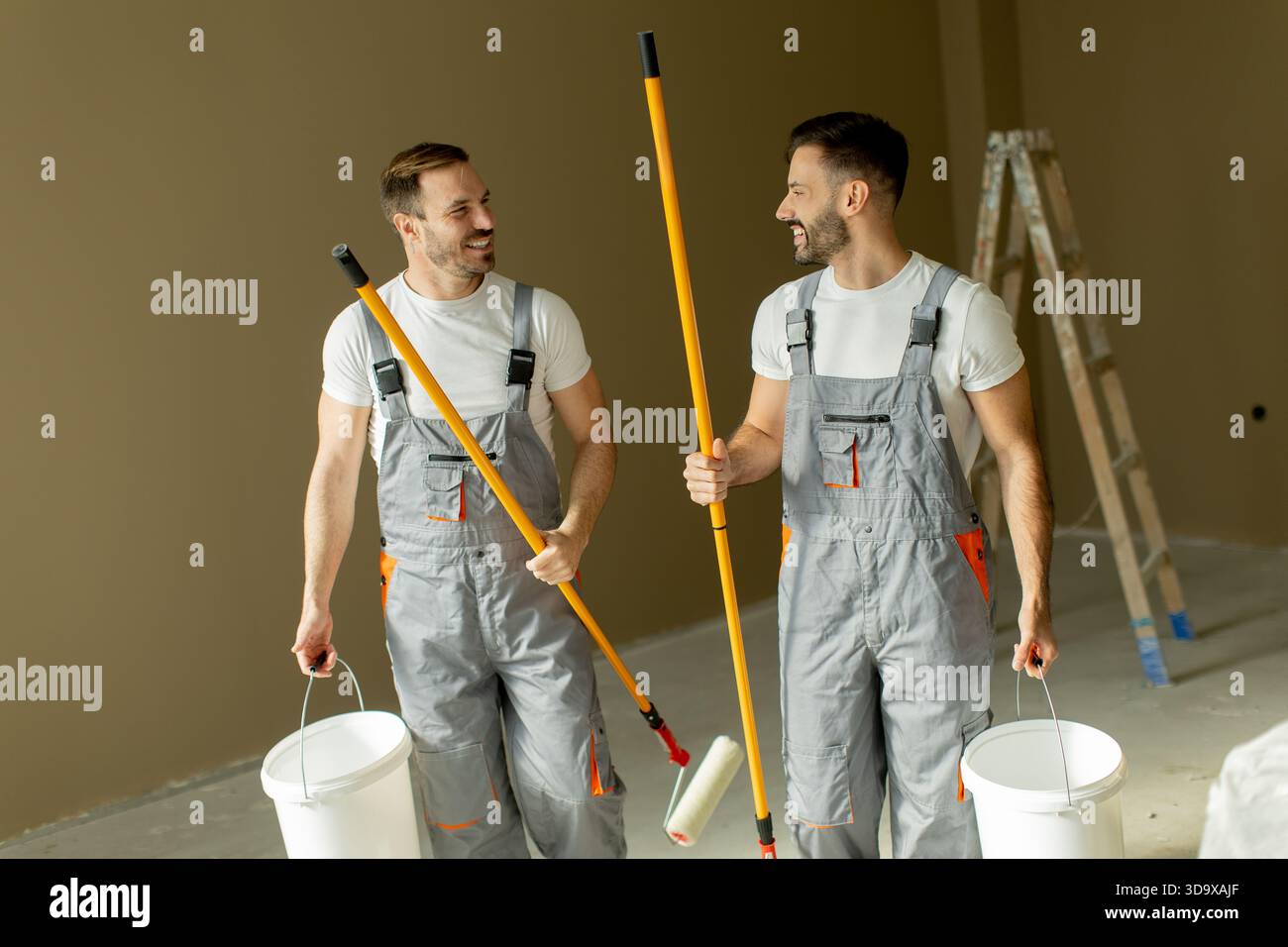 Zwei Männer lächeln und plaudern, während sie Malerwalzen und Eimer in einem leeren Raum halten. Sie bereiten sich darauf vor, die Wände zu streichen und Teamwork zu zeigen Stockfoto