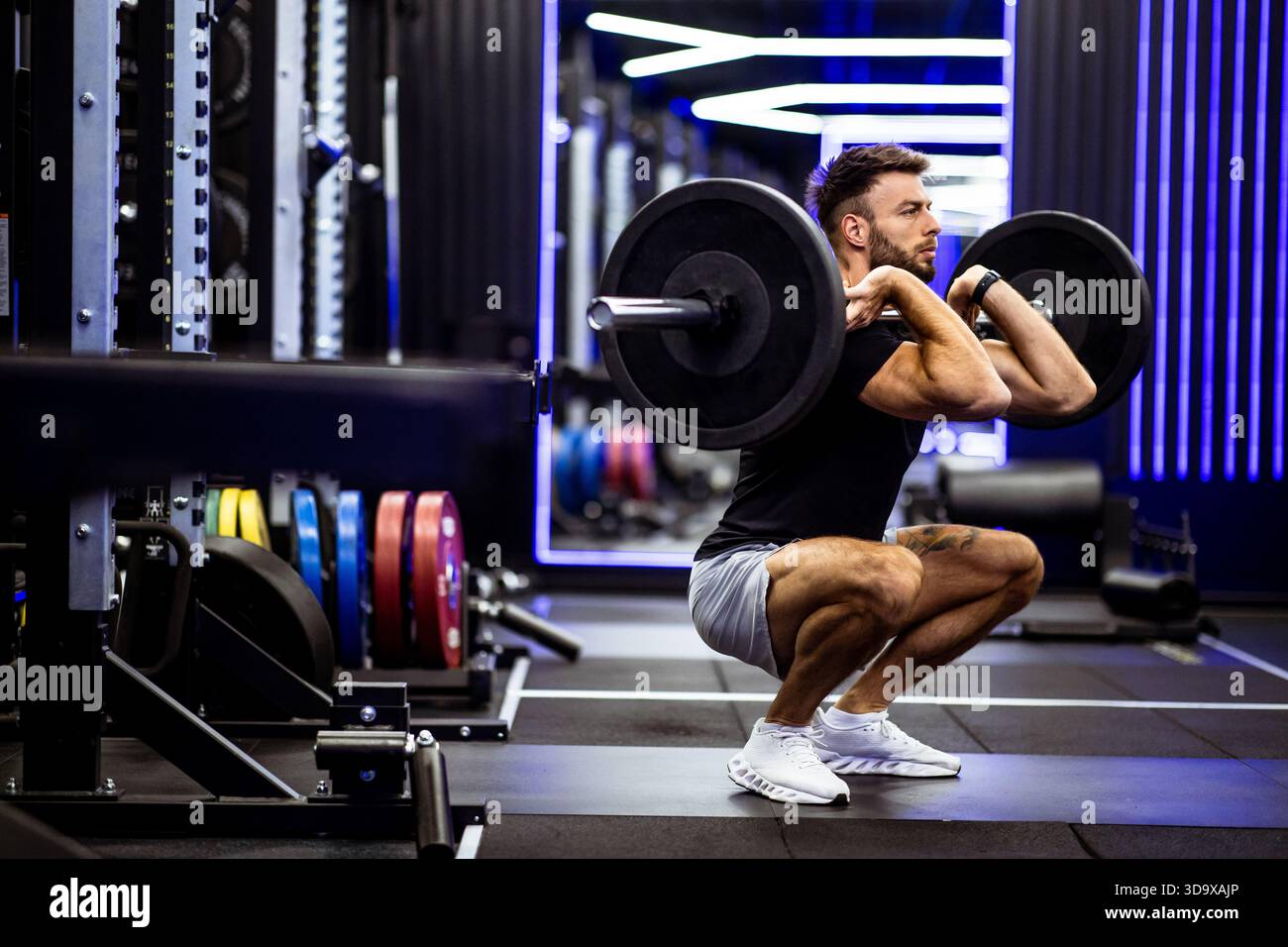 Ein Mann spielt eine Langhantelhocke in einem modernen Fitnessstudio mit lebhafter Beleuchtung. Er konzentriert sich intensiv auf sein Workout, umgeben von verschiedenen Gewichtsplatten und Stockfoto